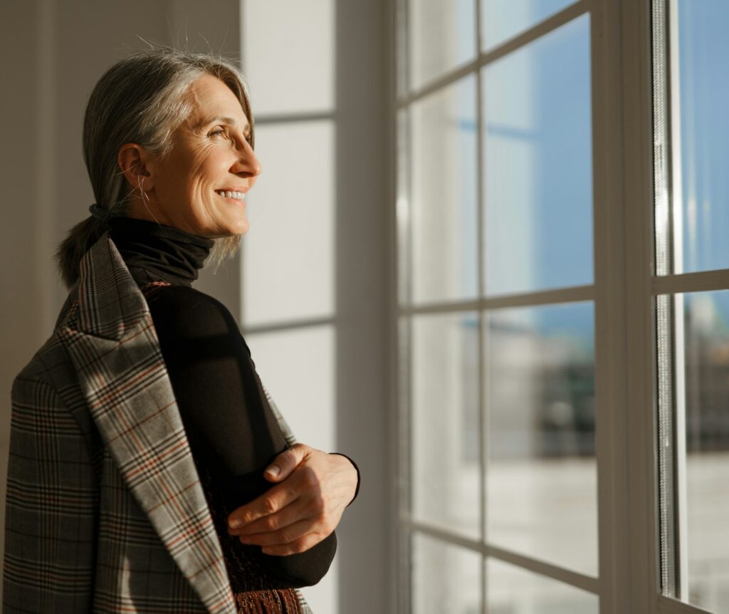 european senior woman with grey hair smiling while looking out window e1722517245943